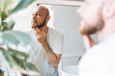 Handsome Bald Man Looking At Mirror And Touching Face In Bathroom