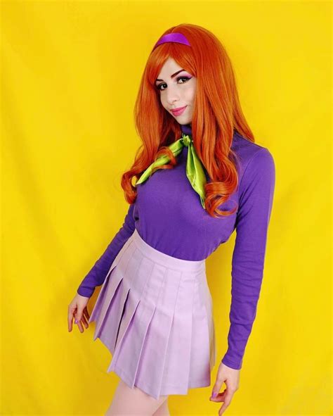 A Woman With Long Red Hair Wearing A Purple Top And Pink Pleated Skirt Posing In Front Of A