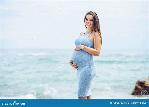 Beautiful Pregnant Woman In A Blue Tight Dress On The Beach Touching Her Belly With Love And