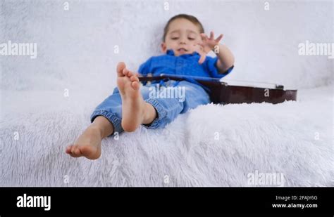 A Child With A Guitar Is Lying On A White Sofa Touching The Strings Of