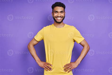 Happy African Man Holding Hands On Hip And Smiling While Standing Against Purple Background