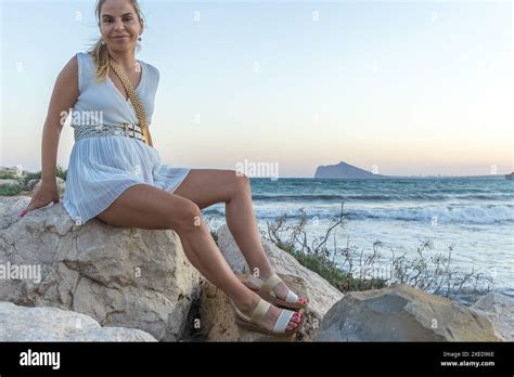 This Image Captures A Serene Moment Of A Beautiful Blonde Woman Seated On Coastal Rocks She Is