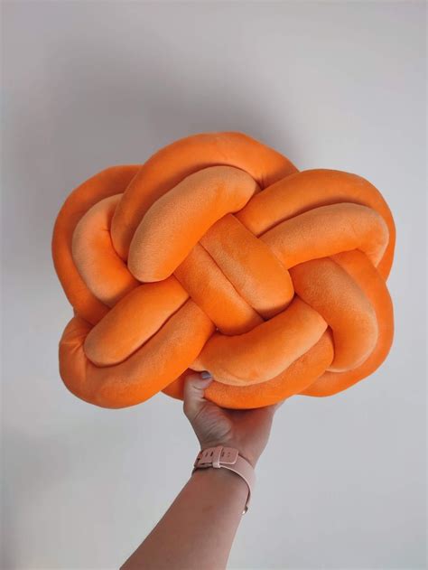 A Hand Holding An Orange Knot Pillow In The Shape Of A Ball On A White