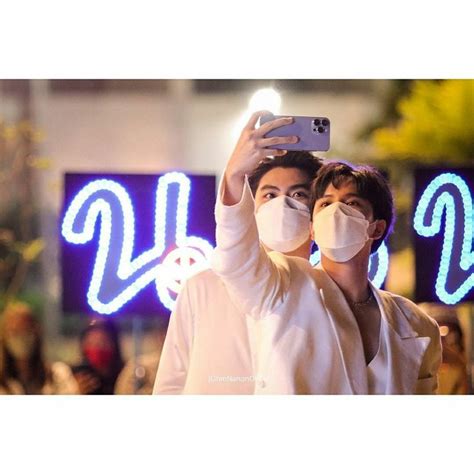 A Man And Woman Wearing Face Masks Taking A Selfie In Front Of Neon Signs