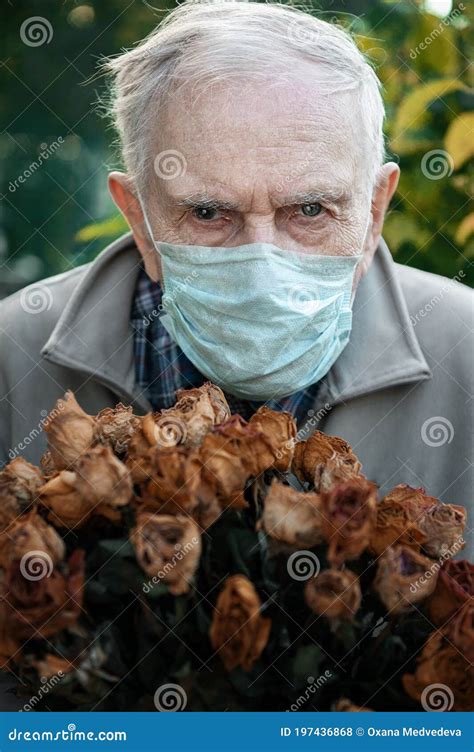 Old Man In A Medical Mask With A Bunch Of Dried Wilted Roses The