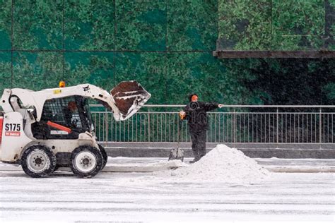 A Utility Worker And A Small Loader Excavator Remove Snow From The Road