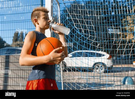 Teenager boy playing street basketball with ball on outdoor city basketball court drinking water ...