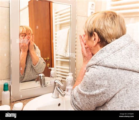 Senior Woman In Bathrobe Touching Her Soft Face Skin Looking In Mirror