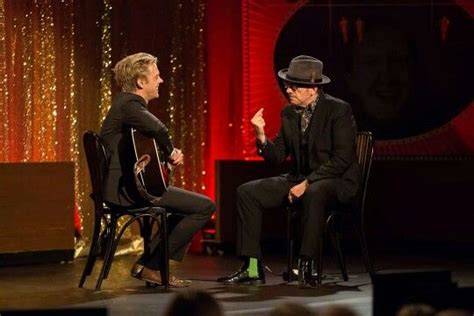 Two Men Sitting On Chairs Talking To Each Other In Front Of A Stage