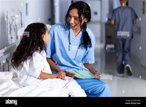 Happy Biracial Female Doctor And Girl Patient Sitting On Bed Talking In