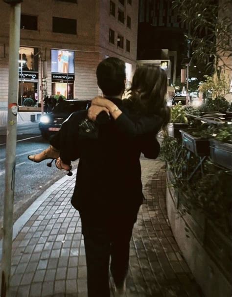 A Man Carrying A Woman On His Back Walking Down The Street At Night