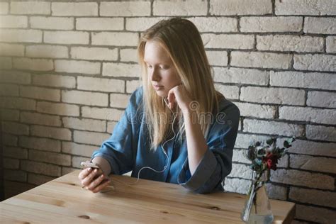 Young Girl In Blue Shirt Sits At Table In Headphones And Thought Stock