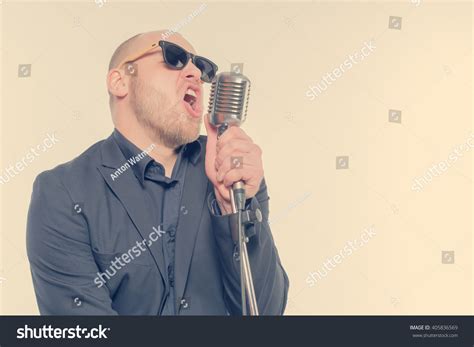 Unshaven Bald Man In A Gray Suit And Sunglasses Holding A Microphone