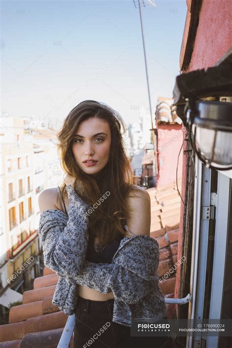 Confident Brunette Girl Posing On Balcony And Looking At Camera With Hand On Locks Person