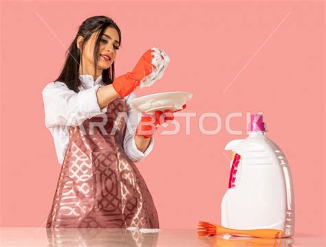 Portrait Of An Arab Woman Wearing Gloves And A Kitchen Apron Washing Dishes With Dish Soap And