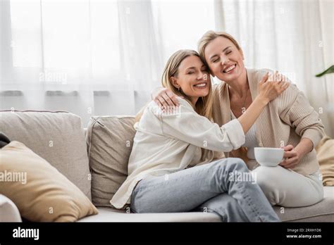 Two Good Looking Blonde Sisters Sitting On Sofa And Hugging With Closed