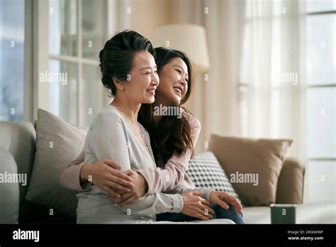 Loving Asian Adult Daughter Sitting On Couch At Home Hugging Senior