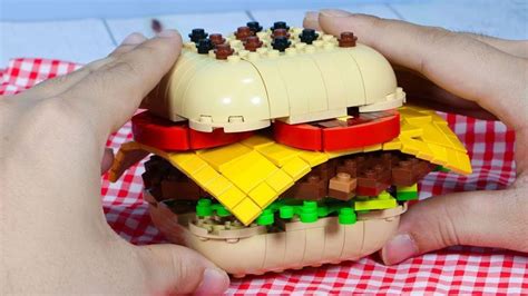 Two Hands Hold A Lego Model Of A Burger On A Picnic Table With Red And