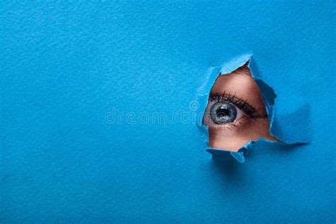 A Female Eye Looks Through A Hole In A Paper Blue Background Stock