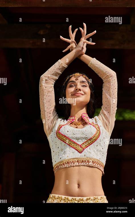 joyful young indian woman in elegant ethnic wear dancing with closed eyes and raised hands ...