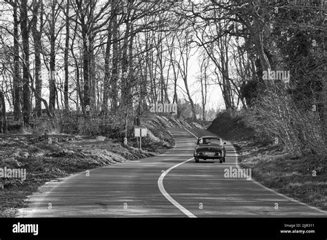 A Grayscale Shot Of An Old Vintage Car Driving Down A Narrow Road