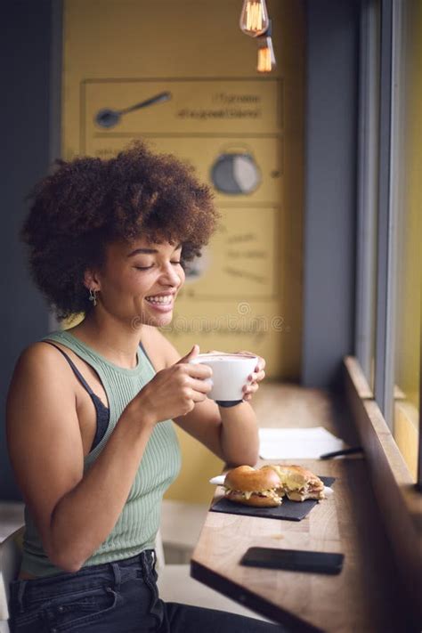 Female Customer In Coffee Shop Window Eating Bagel Sandwich For Lunch