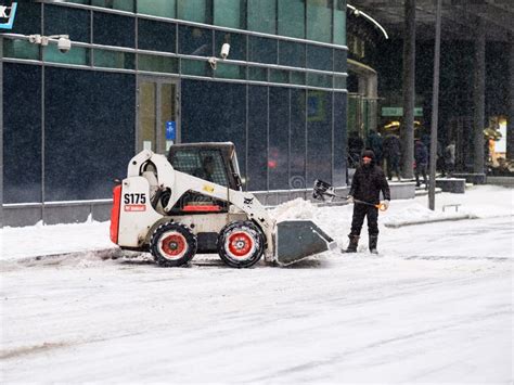 A Utility Worker And A Small Loader Excavator Remove Snow From The Road