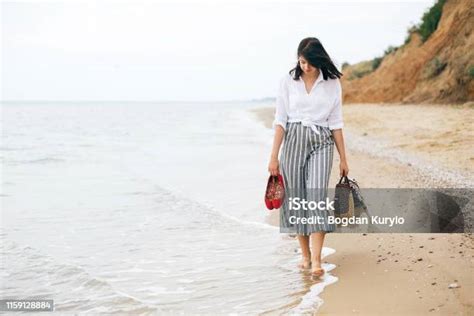 해변에서 맨발로 걷고 가방과 신발을 손에 들고있는 세련된 힙스터 소녀 열대 섬에서 산책을 즐기며 바다에서 휴식을 취하는 행복한