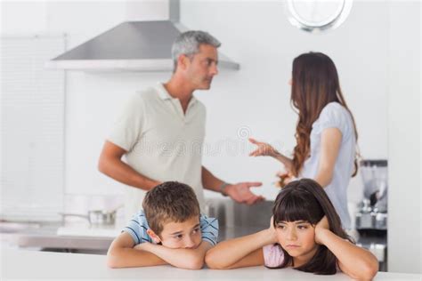 Unhappy Siblings Sitting In Kitchen With Their Parents Who Are F Stock