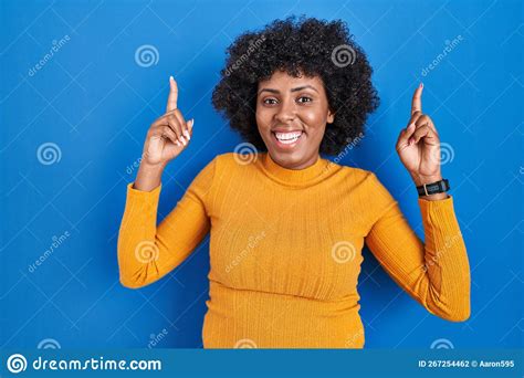 Black Woman With Curly Hair Standing Over Blue Background Smiling Amazed And Surprised And