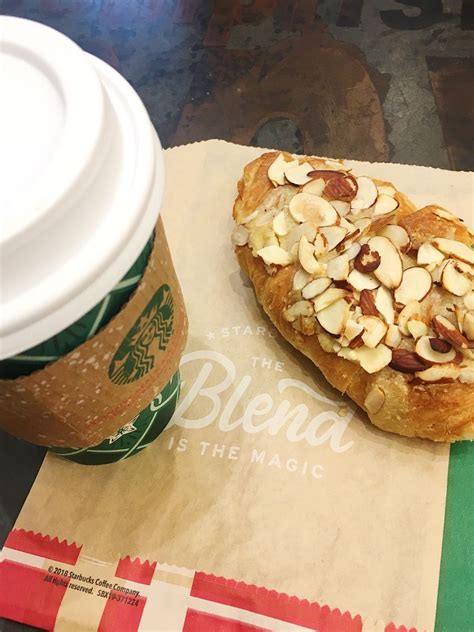 A Pastry With Almonds On It Sitting Next To A Cup Of Coffee And Paper Bag