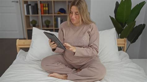 Pregnant Blonde Woman Sitting On A Bed In A Cozy Bedroom Using A Tablet
