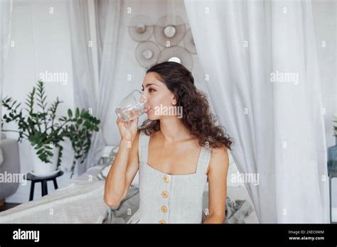 A Charming Curly Haired Brunette In A Linen Sundress Holds A Transparent Cup And Drinks Water