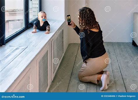 Baby With Pacifier Crawls On Windowsill And Woman Takes Photos Stock