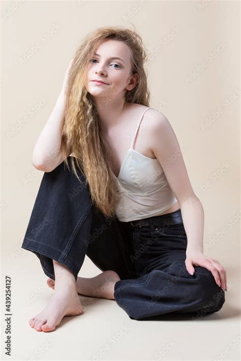 Young Calm Caucasian Blond Girl In White Bra With Jeans Sitting With Folded Legs Over Beige