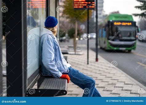 Millennial Guy Sitting On Bench At Bus Stop Looking At Arriving Bus