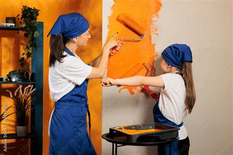 Mother And Girl Painting House Walls With Orange Color Paint Using