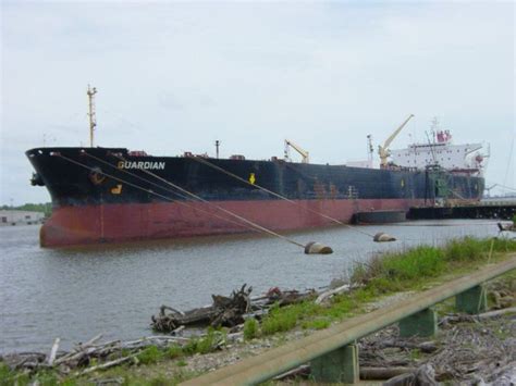 A Large Cargo Ship Is Docked In The Water Next To Some Logs And Grass On The Shore