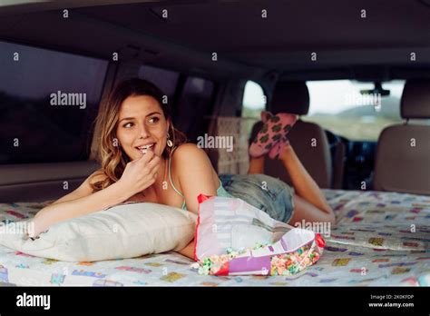 Blonde Cheerful Pretty Woman Lying On Folding Bed Inside A Minivan While Eating Popcorn Stock