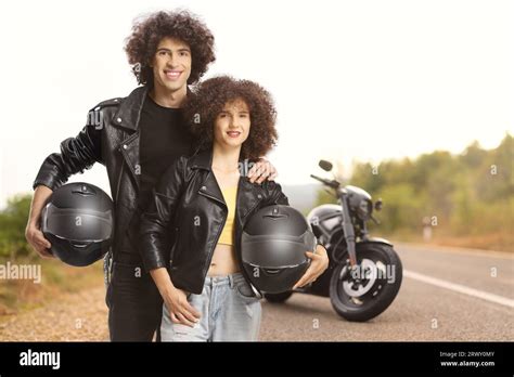 Couple In Leather Jackets Holding Helmets In Front Of A Parked