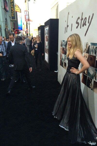 A Woman In A Long Black Dress Standing Next To A Wall With Pictures On It