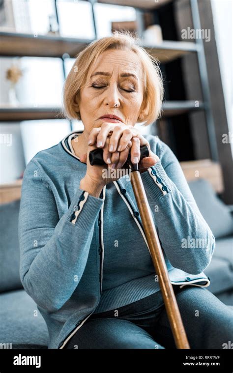 Sad Senior Woman With Eyes Closed Leaning On Walking Stick At Home