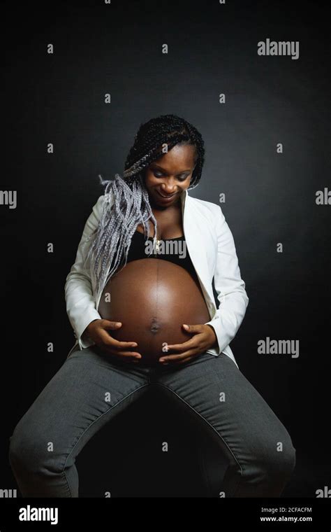 Young Pregnant African American Female With Braids Wearing White Blazer And Jeans Stroking Belly