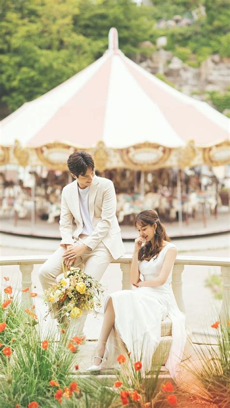 A Man And Woman Sitting On A Bench In Front Of A Carousel With Red Flowers