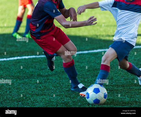 Boys In Red And Blue Sportswear Plays Football On Field Dribbles Ball