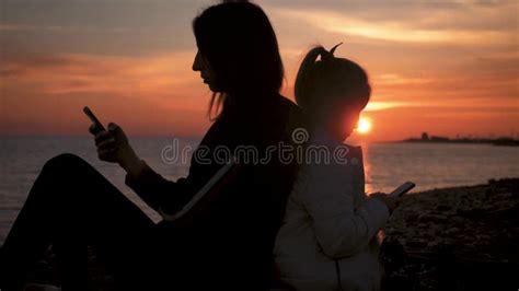 Silhouettes Mother And Daughter Use Mobile Phone On The Beach At Sunset