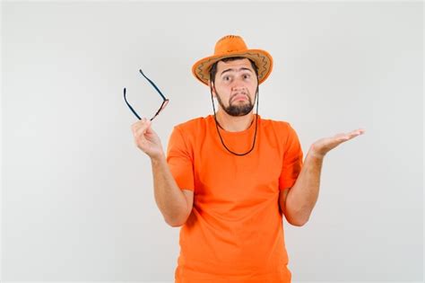 Jovem macho segurando óculos um gesto indefeso em t shirt laranja