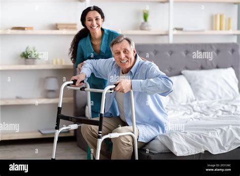 Female Nurse Helping Elderly Male With Walking Frame Stand Up From Bed