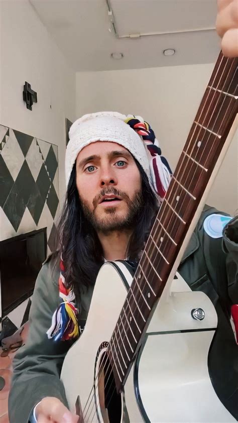 A Man With Long Hair Is Holding A Guitar In His Right Hand And Looking Up At The Camera