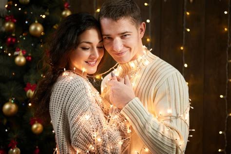 Young Couple Together In Christmas Lights And Decoration Dressed In White Fir Tree On Dark